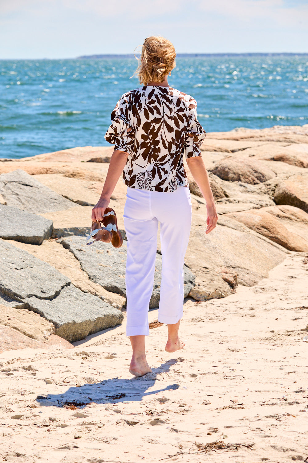 Woman in a patterned top and white pants standing on a rocky beach.