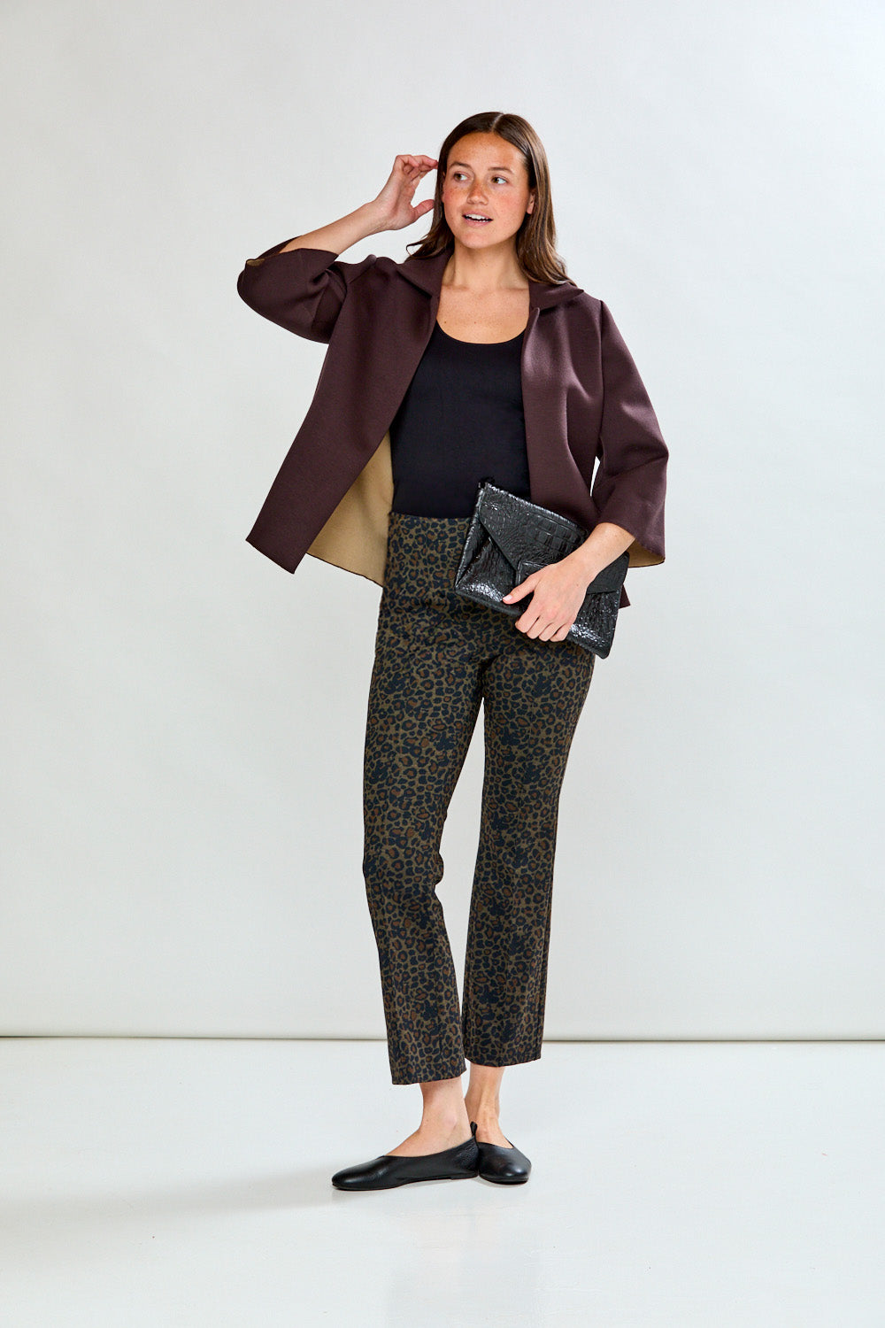Woman in a brown blazer and black pants holding a clutch against a white background