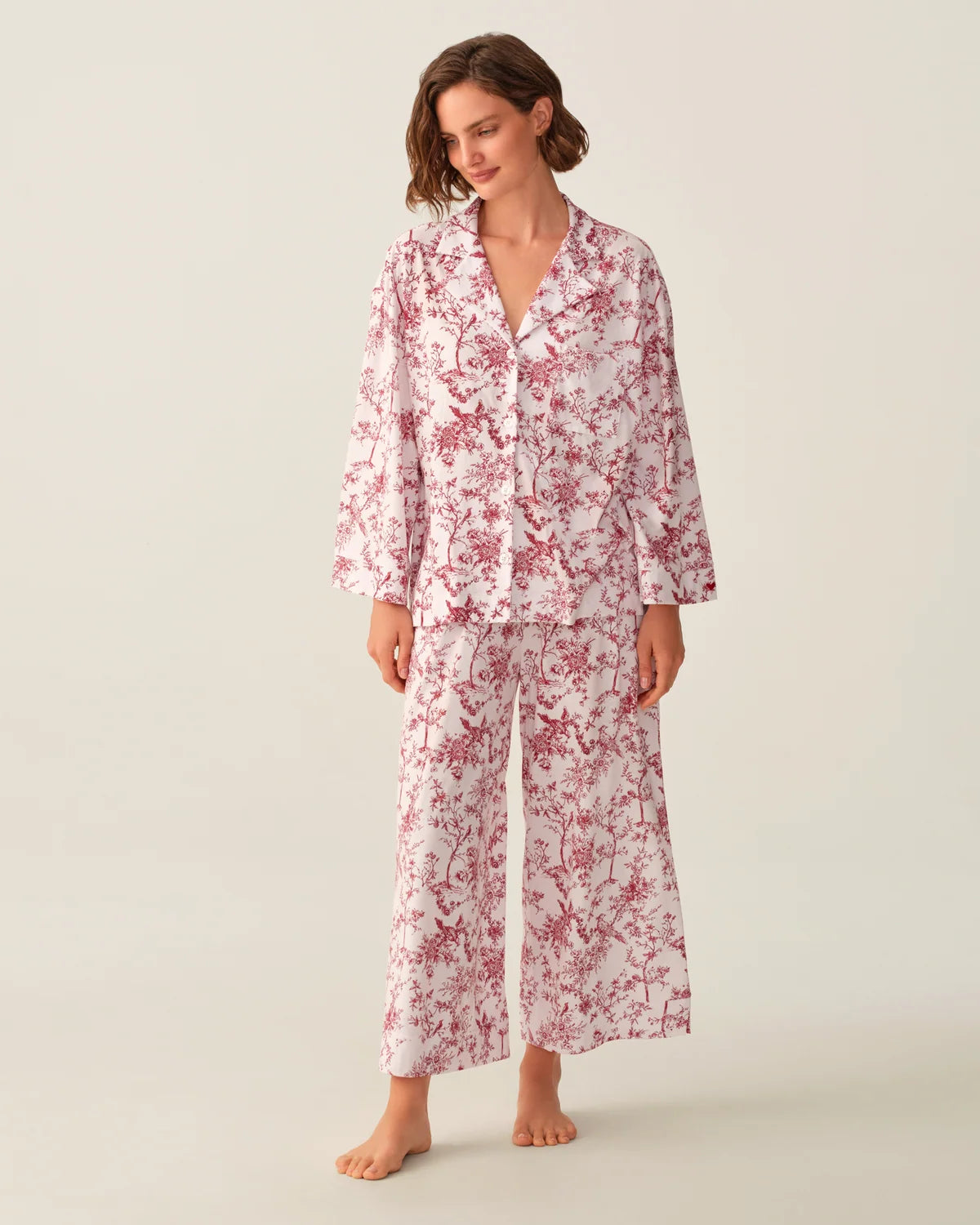 A woman modeling a matching pajama set with a white base and red floral print, consisting of a long-sleeve button-up top and wide-leg pants, against a plain light beige studio background.