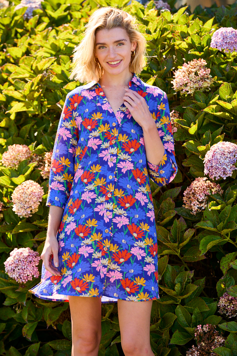 Woman wearing a colorful floral dress standing in front of hydrangea plants.