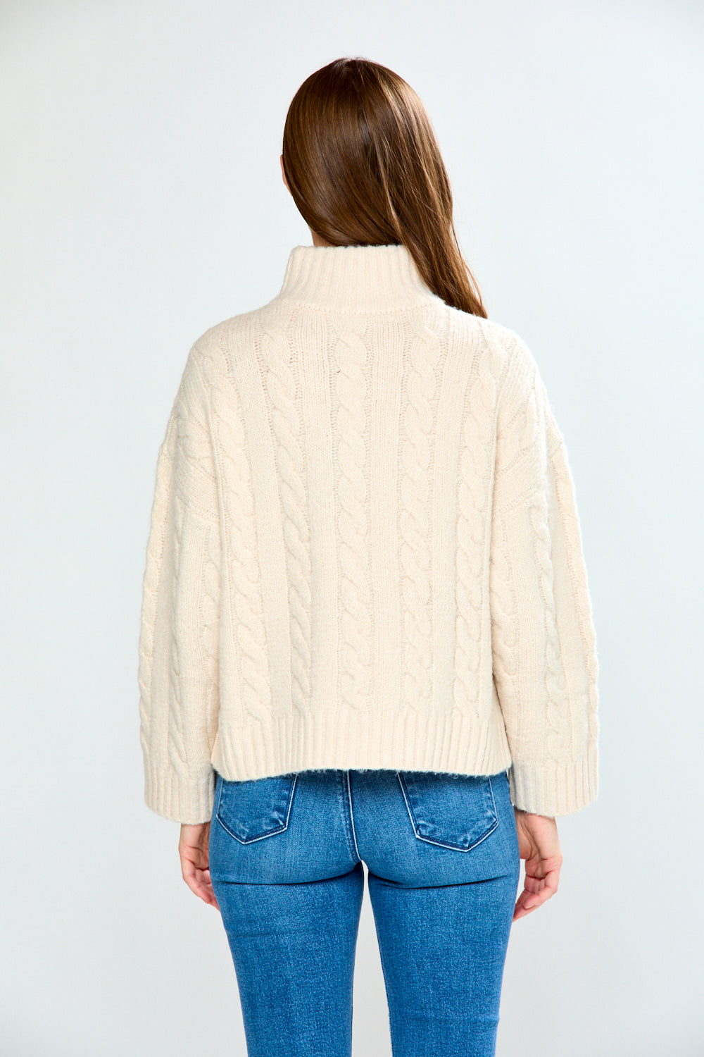 Woman wearing a oatmeal cable knit sweater and blue jeans on a white background