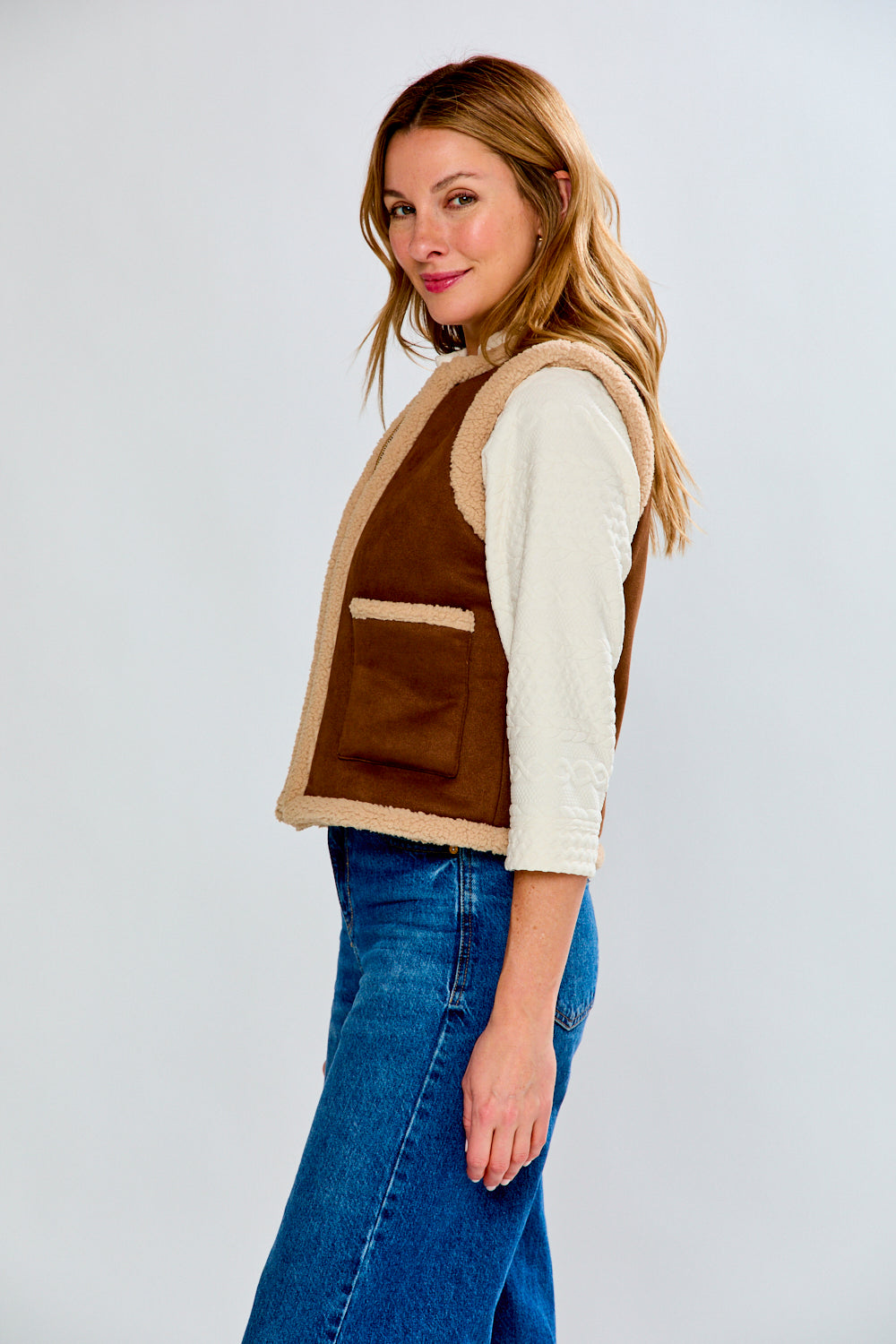 Woman wearing a brown vest over a white shirt with blue jeans on a light gray background