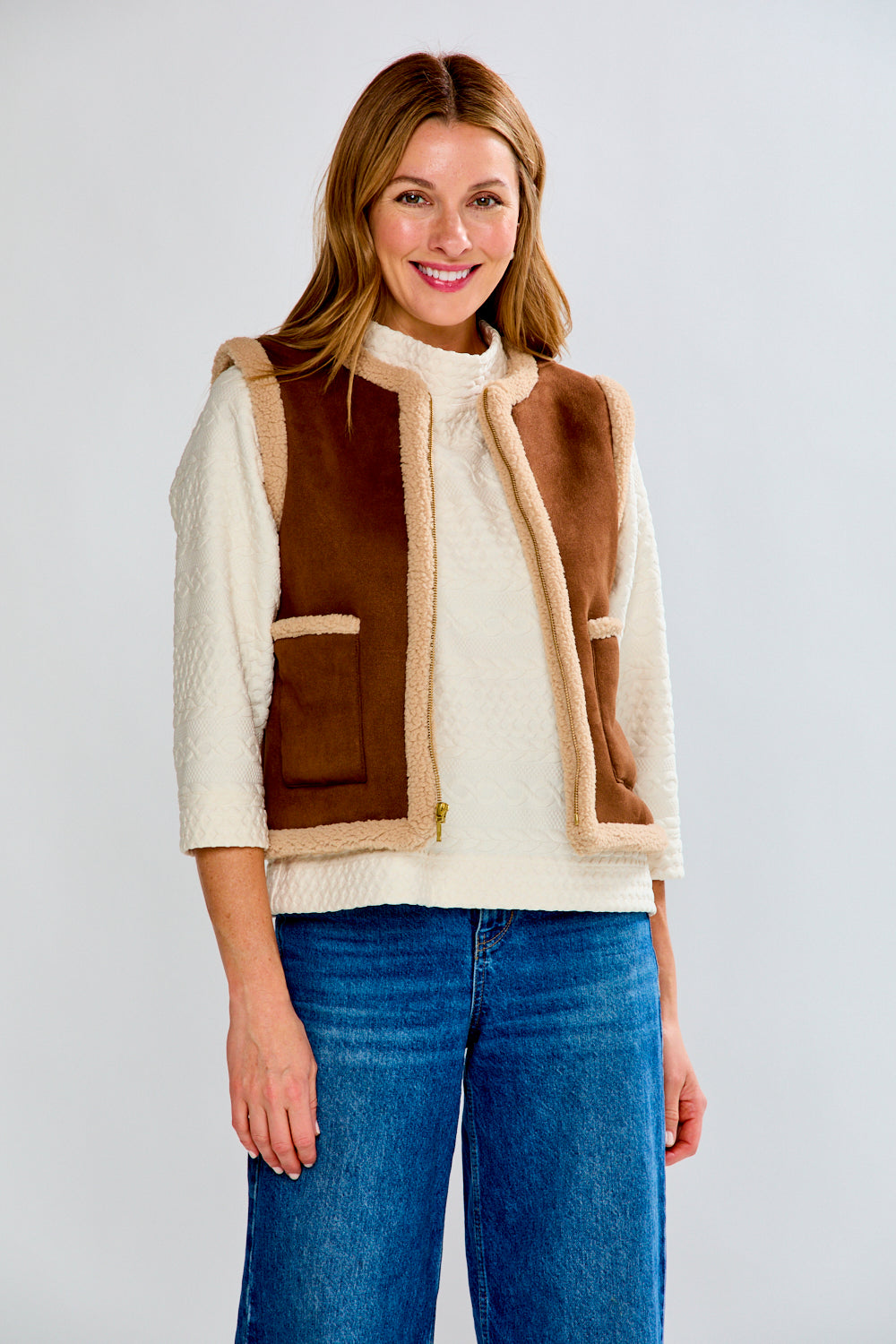 Woman wearing a brown and beige vest over a white shirt with blue jeans on a light gray background