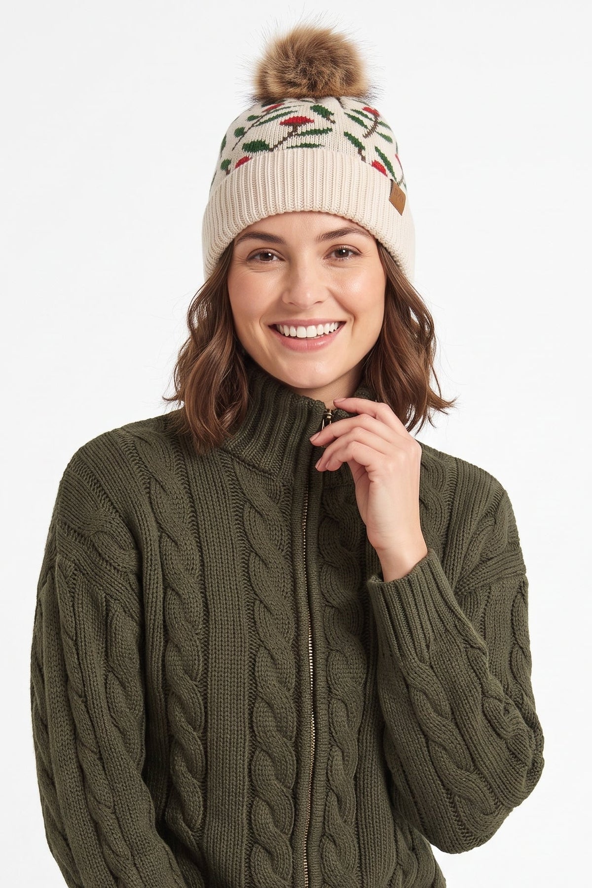 A woman wearing an olive green cable-knit mock neck sweater, a beige knit beanie with a brown pom-pom and colorful floral pattern, and light blue jeans, posed against a solid white background.