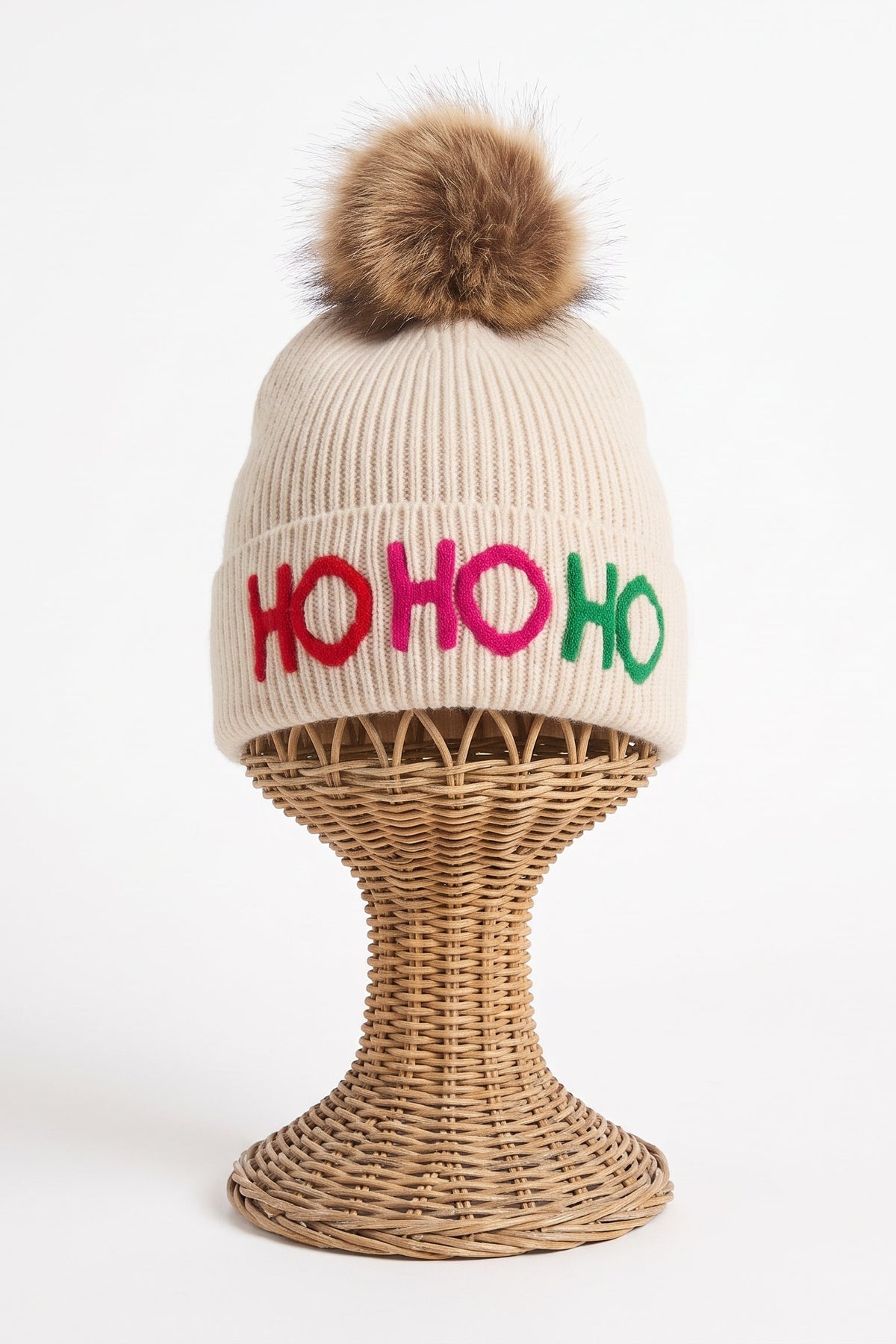 A ribbed knit beanie hat in warm cream with a brown faux-fur pom-pom on top and colorful embroidered 'HO HO HO' lettering across the front. The hat is displayed on a woven wicker stand against a clean white background.