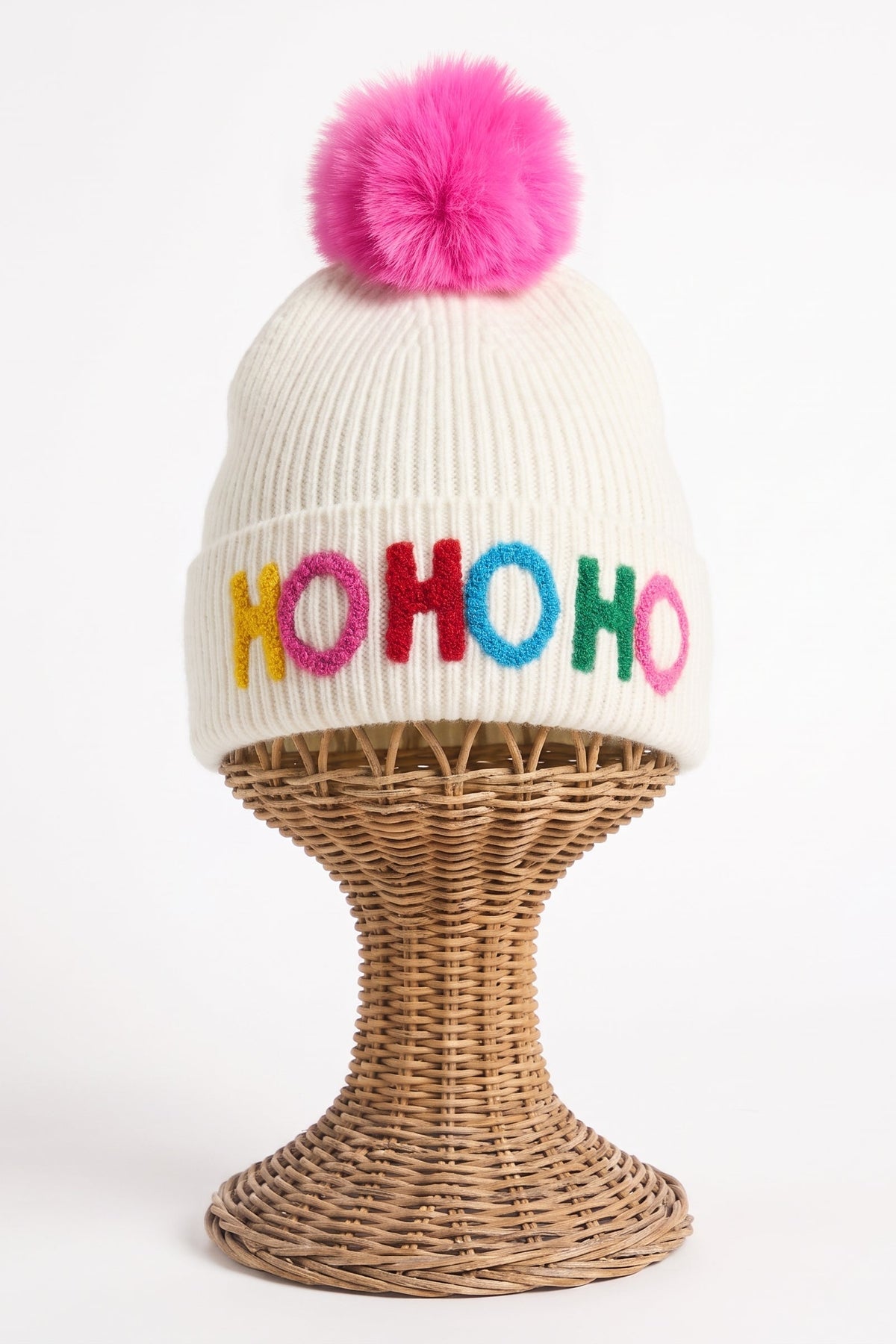 An off-white ribbed knit beanie with a bright pink pom-pom on top and colorful embroidered 'HOHOHO' lettering on the front, displayed on a woven wicker stand against a white background.