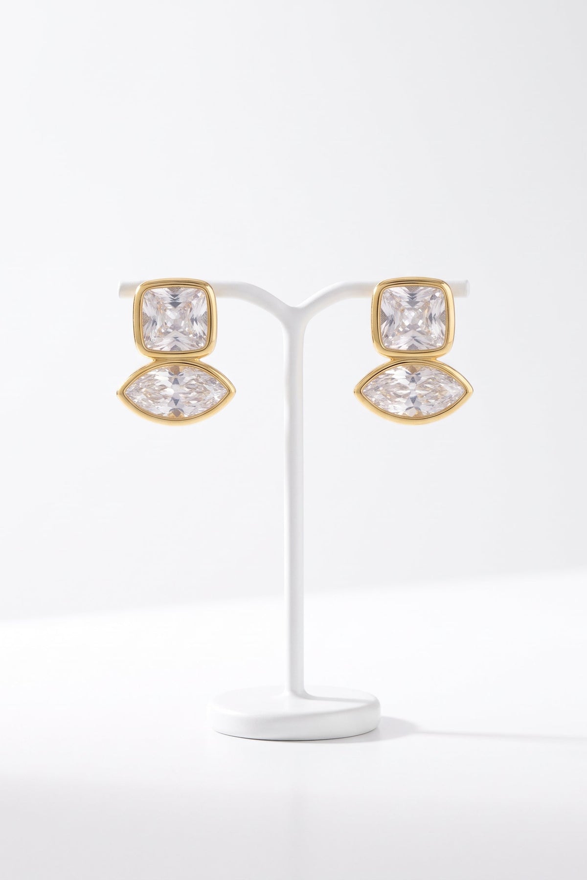 A pair of gold stud earrings with clear marquise-cut and cushion-cut gemstones, displayed on a white jewelry stand against a clean white background.