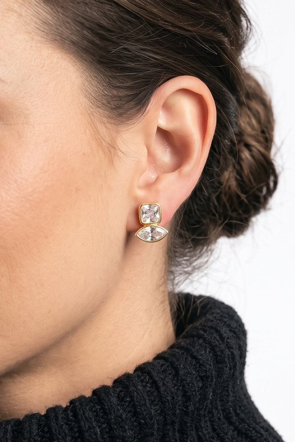 A close-up of a woman wearing a small silver-toned dangle earring with clear stones, paired with a black ribbed turtleneck sweater against a plain white background.