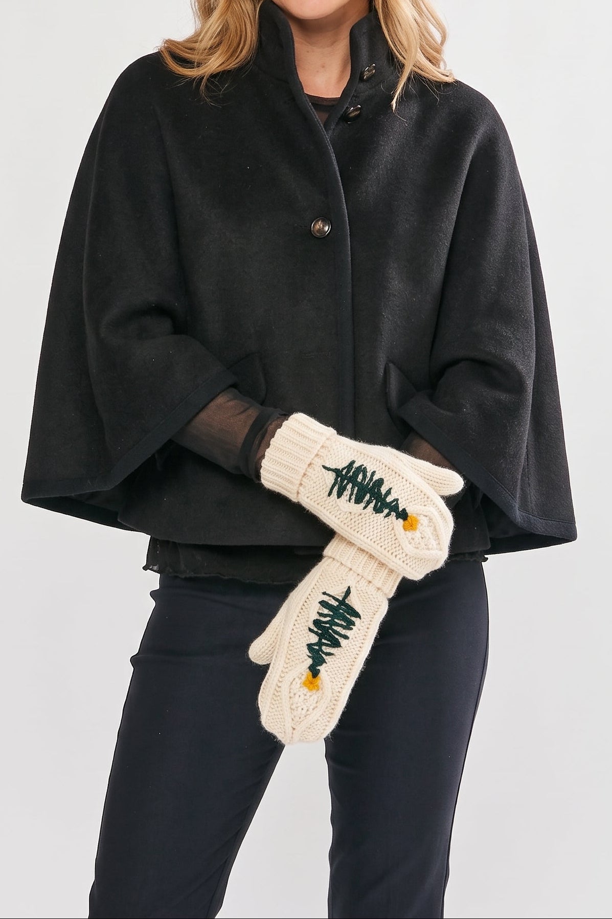 A woman wearing a black cape-style coat and cream knit mittens with green pine tree embroidery, standing against a light gray studio background.