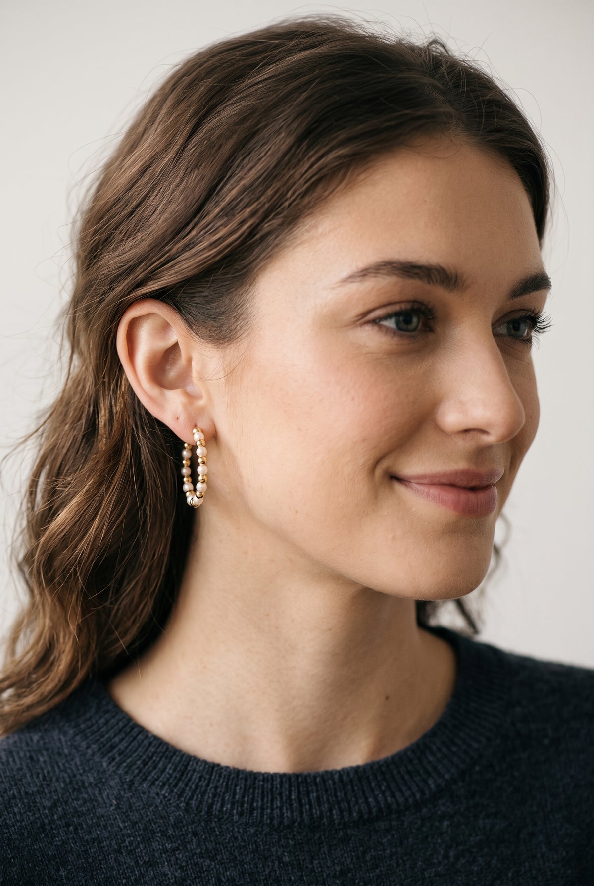 A woman with brown hair wearing gold hoop earrings with small white bead details, shown in a close-up profile against a light gray background.