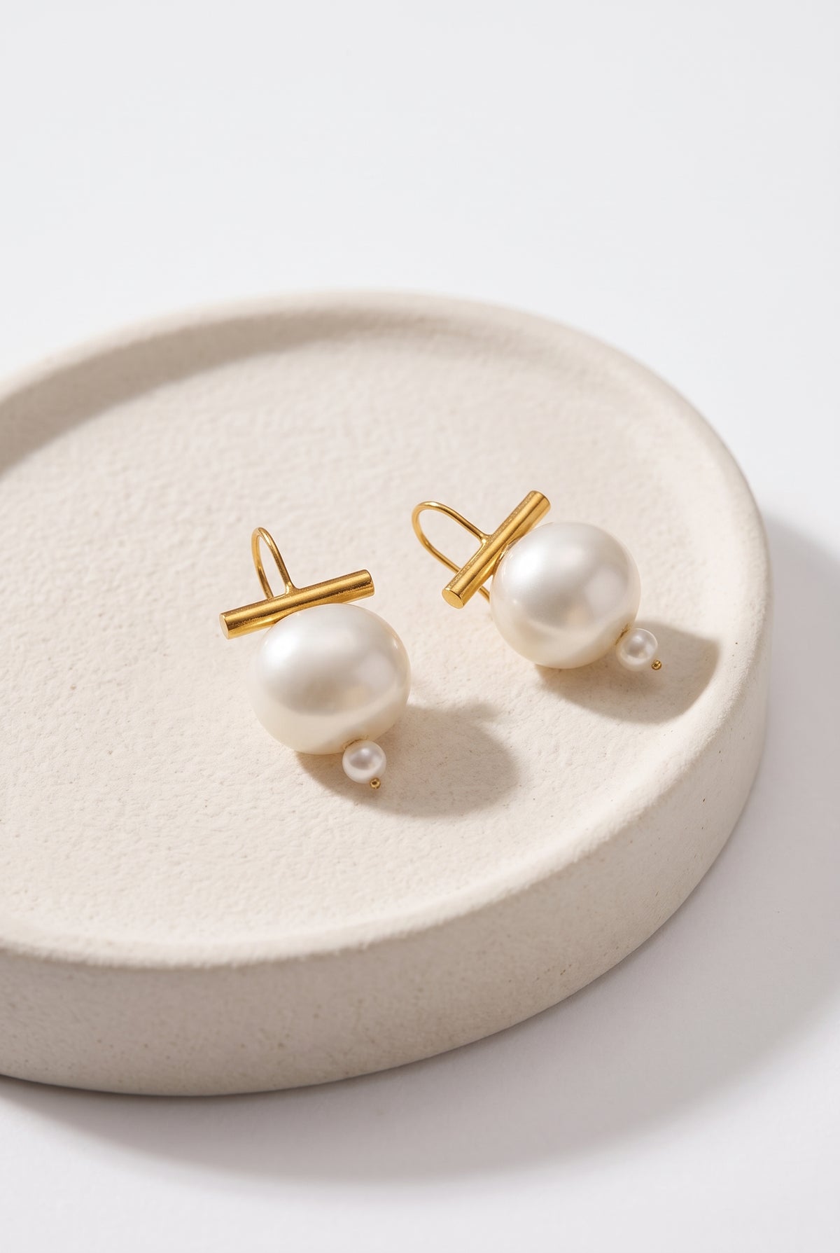 A pair of gold and pearl stud earrings displayed on a round beige ceramic dish against a clean white background.