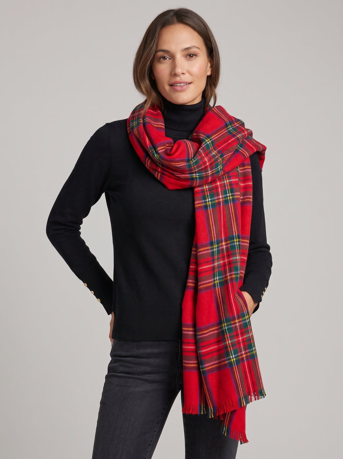 A woman wearing a classic red tartan plaid scarf with green, yellow, and blue stripes, styled over a black turtleneck sweater and dark jeans against a solid light gray studio background.