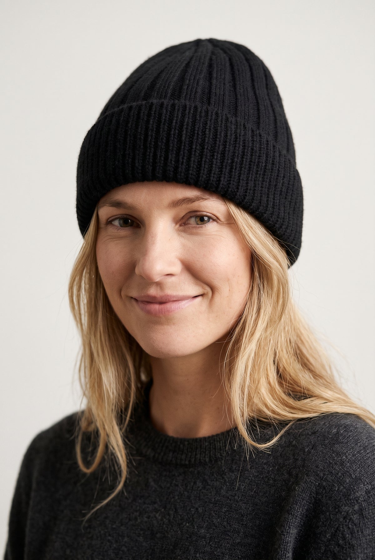 A woman with blonde hair wearing a black ribbed knit beanie and a dark gray knit sweater, photographed against a light gray background.