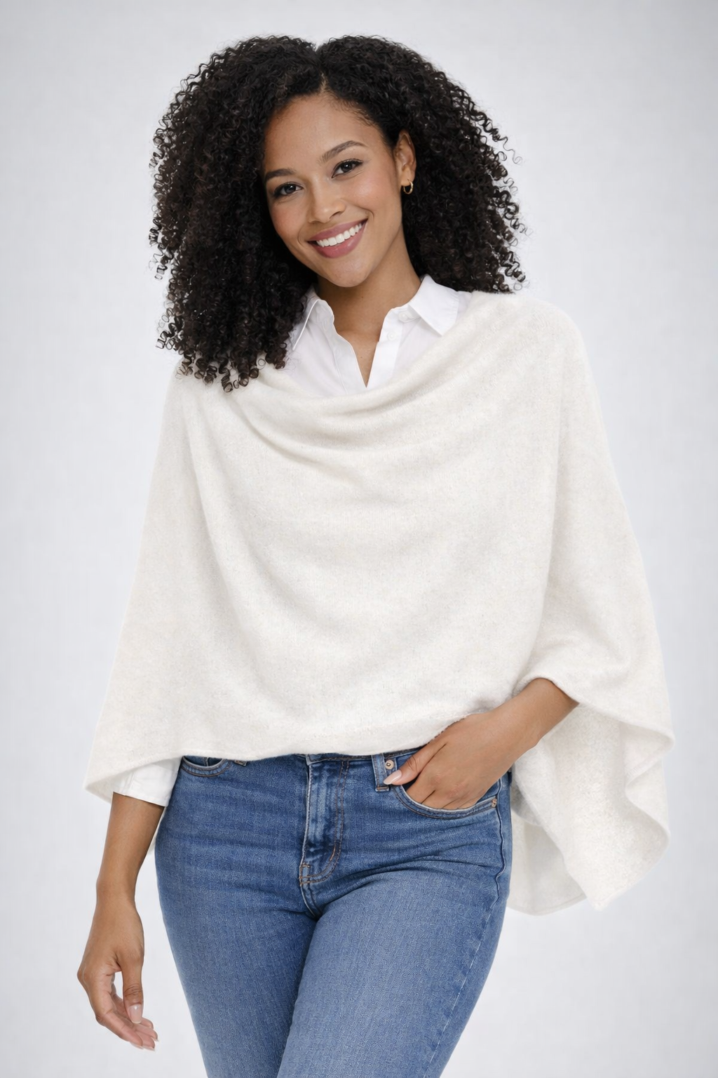 Woman wearing a white poncho over a white shirt with blue jeans on a plain background